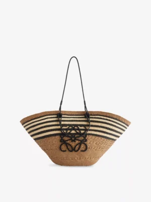 A wide-brimmed woven straw tote bag inspired by the Loewe Ola Medium Embellished Leather Shoulder Bag, featuring black and white horizontal stripes near the top, black leather handles, and a large black decorative logo on the front.