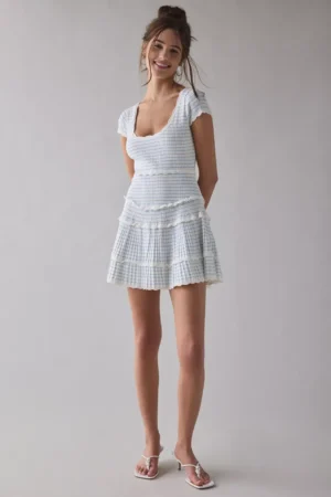 A woman stands against a plain gray background wearing a fitted white top and the LoveShackFancy Sydow Dress, a short, tiered, ruffled skirt with a light blue plaid pattern. She completes the look with white sandals.