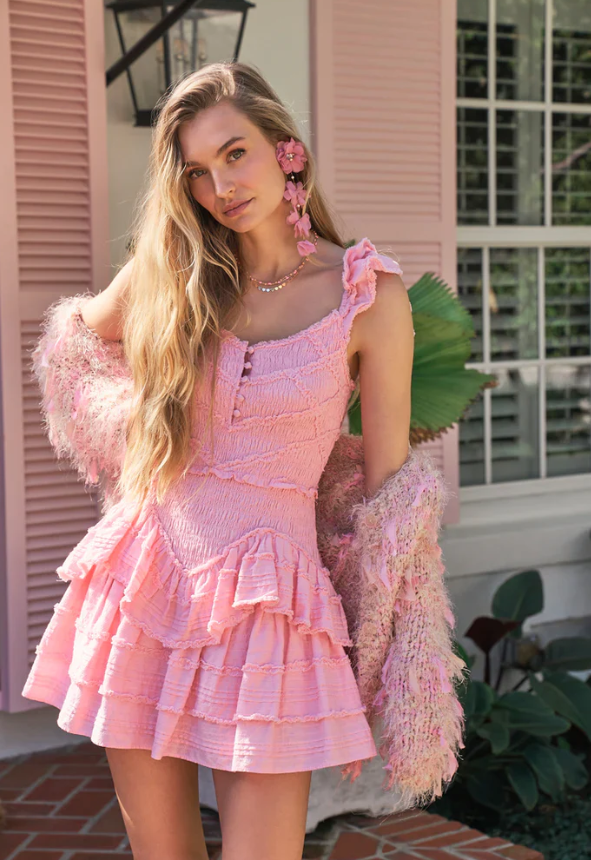 A woman wearing a light pink ruffled dress—perfect for those who love pink summer dresses—and a textured shawl stands outdoors in front of a window with white shutters. She has long blonde hair, flower-shaped earrings, and a necklace.