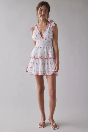 A woman stands against a plain background wearing the LoveShackFancy Docila Heritage Lace-Trim Cotton Mini Dress, featuring red floral embroidery, scalloped edges, and white sandals. Her hair is styled in a loose updo.