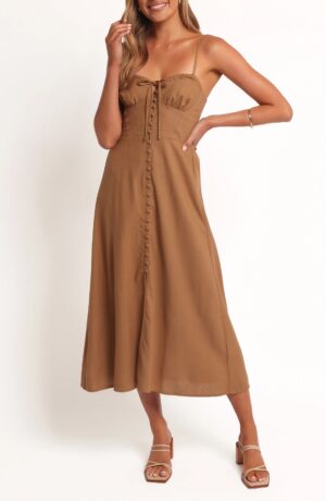 A woman stands against a white background wearing the Petal & Pup Velda Floral Cotton Midi Dress, featuring a tan, sleeveless, button-front style with spaghetti straps and a front tie. She completes the look with heeled sandals and a bracelet.