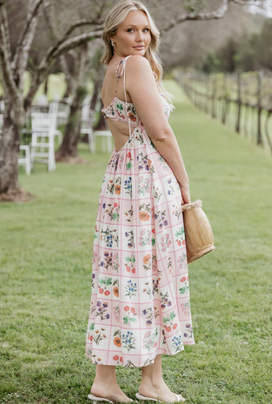 A woman stands on grass wearing a flowy beach dress with a grid of colorful floral patterns. She holds a wicker handbag and looks over her shoulder, with trees and chairs in the background.