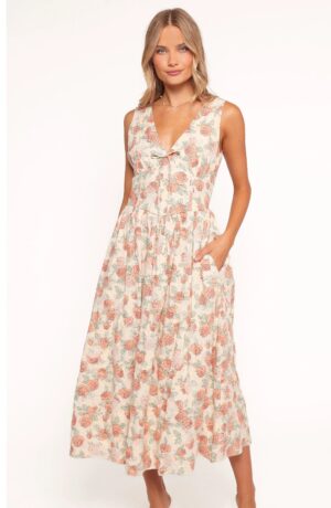 A woman stands against a plain background wearing the Petal & Pup Andreya Midi Dress, a sleeveless, V-neck, ankle-length piece featuring a floral pattern in soft pink and green tones. She has her hands in the dress pockets and is looking at the camera.