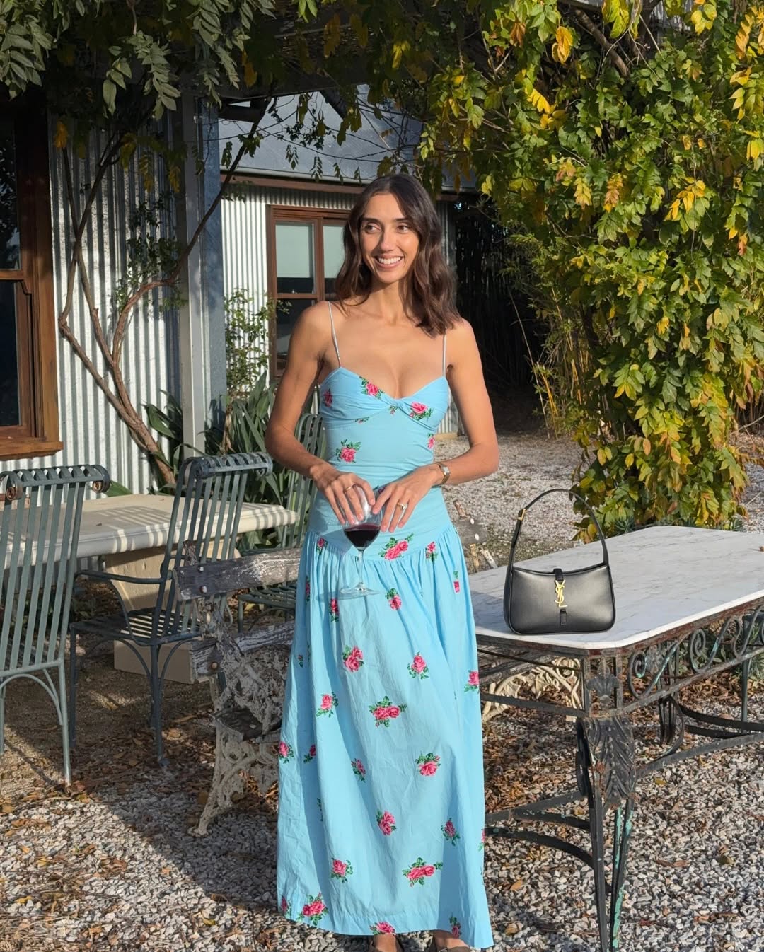 A woman in a light blue, floral-patterned dress stands outdoors by a white metal table and chairs, holding a glass—perfect inspiration for picnic dress ideas. A black handbag sits on the table, with greenery and a building in the background.