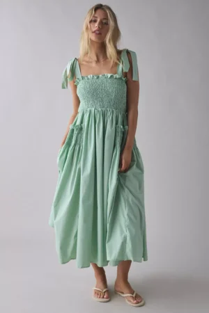 A woman stands against a plain background wearing the Sister Jane Forget Me Not Beaded Floral Appliqué Asymmetric Midi Dress with tie straps, a smocked bodice, and gathered skirt. She pairs it with flat sandals and her loose, blonde hair.