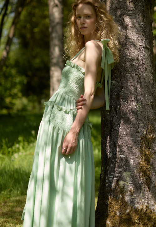 A woman with long curly hair stands outdoors, leaning against a tree. She is wearing a light green, sleeveless, flowing Summer Baby Shower Dress. The sunlit, green background suggests a natural, park-like setting.