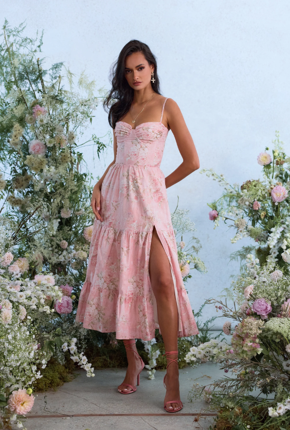A woman wearing a pink floral Hampton's wedding guest dress with a high slit stands among green plants and flowers. She has long dark hair, pink heels, and poses with one hand on her hip against a light background.