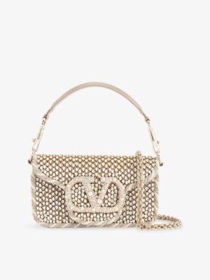 A small beige Valentino Garavani VLogo Floral Embroidered Bucket Bag covered with pearls, featuring a large embellished V-shaped logo on the front, a short handle, and a gold chain shoulder strap.