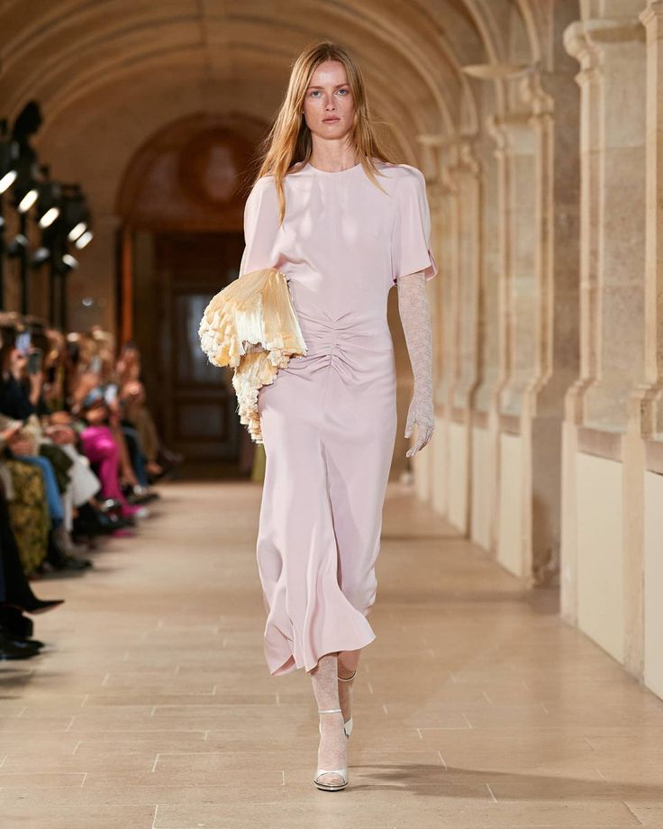 A model walks down a stone hallway runway in a light pink, ruched, mid-length dress with sheer gloves and white open-toe heels, embracing 2025 fashion trends. She holds a beige ruffled accessory as the audience admires pink summer dresses from the side.