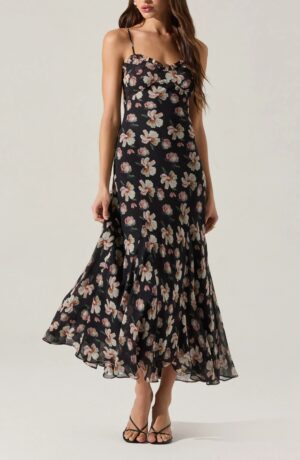 A woman wearing a sleeveless, ankle-length black dress with a floral print and thin straps, reminiscent of the ASTR the Label Melani Strapless Minidress, stands against a plain background. She pairs it with chic black strappy heels.