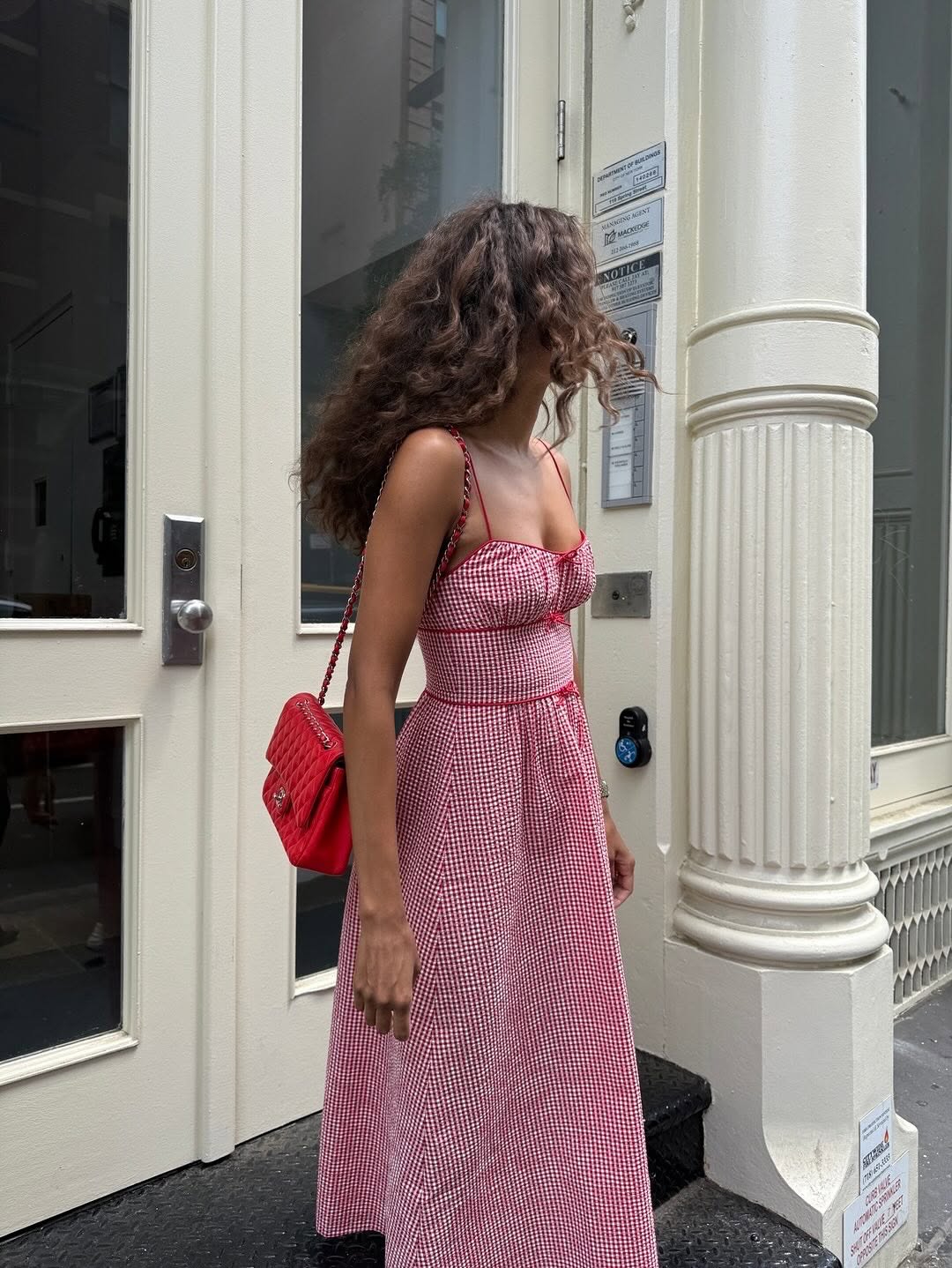 A person with long, curly hair wearing a red and white checkered Doen Summer Dress and carrying a red shoulder bag stands next to a white building with columns, looking away from the camera.