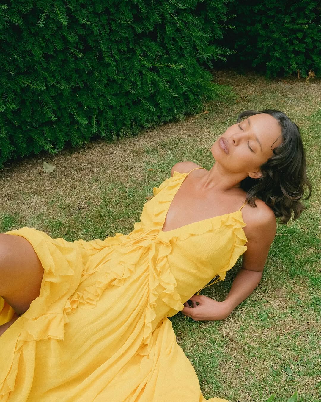 A woman in a bright yellow Doen Summer Dress sits on the grass with her eyes closed, leaning back on one arm. Green shrubbery forms a lush backdrop.