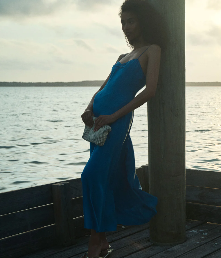 A person in a sleeveless blue Doen Summer Dress stands on a wooden dock by the water, holding a white clutch and leaning against a vertical post under a cloudy sky.