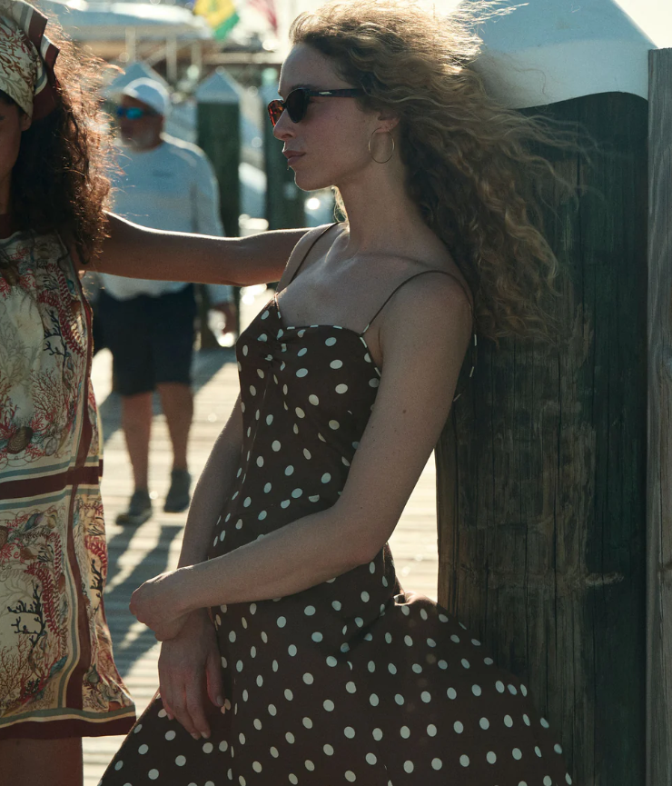 A woman in a brown polka dot dress, reminiscent of Doen Summer Dresses, and sunglasses leans against a wooden post on a dock. Her curly hair blows in the wind as another person in a patterned outfit stands partially visible beside her.