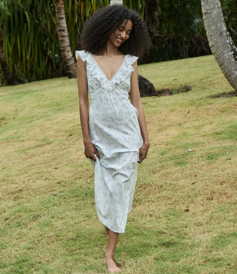 A woman with curly hair stands barefoot on grass, wearing a light-colored, sleeveless, ruffled Summer Baby Shower Dress with a floral pattern. Palm trees and greenery are visible in the background.