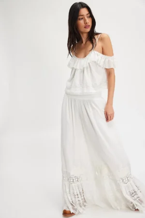 A woman stands slightly angled, wearing the Free People Merrick Midi Dress—a white off-the-shoulder top with ruffles and a matching long, flowing lace skirt. The outfit features lace details at the hem and waistband against a plain white background.