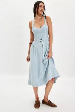 A woman stands against a plain white background, wearing a light blue dress inspired by the Free People Morning Meadows Long-Sleeve Maxi Dress. It features a sweetheart neckline, belted waist, brown lace-up shoes, long dark hair, and a beaded necklace.