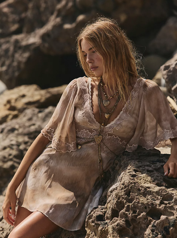 A woman with long, wavy blonde hair stands among large rocks, wearing a light brown, lace-trimmed cute mini dress and layered necklaces with heart pendants. She looks downward and slightly to the side.
