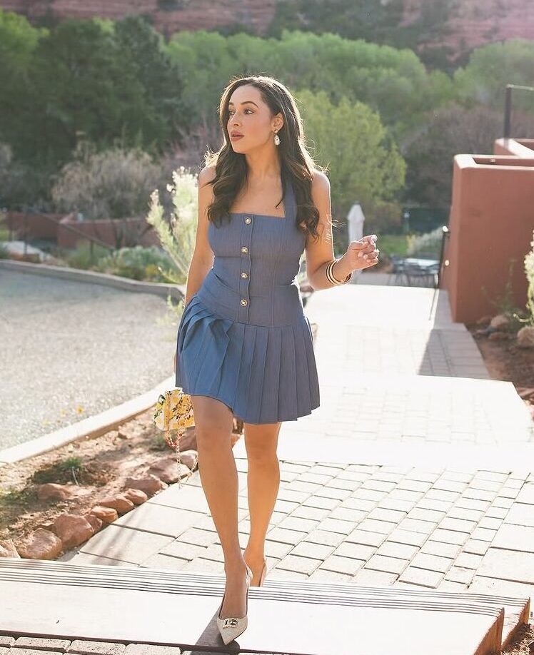 A woman in a cute mini dress and white heels walks on a paved path outdoors, sunlight streaming in from the upper right, with red rock cliffs and green trees in the background.