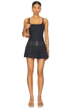 A woman stands facing forward wearing a dark denim mini dress with wide straps, a belted waist, and side pocket detail reminiscent of the Lioness District Maxi Dress. She carries a shoulder bag and wears tan heeled sandals. Her hair is tied back.