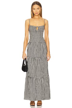 A woman stands facing forward, wearing a sleeveless, black and white gingham maxi dress with a small front cutout and tiered skirt—reminiscent of the More To Come Margo Tie Strap Mini Dress. She holds a small black handbag and wears flat sandals.
