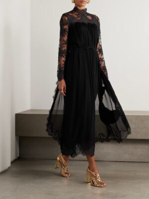A person wearing a Zimmermann Hypnotic Picnic Belted Lace-Trimmed Linen and Silk-Blend Midi Dress, gold earrings, and gold strappy high-heeled sandals stands indoors near a concrete bench.
