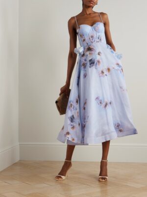 A woman stands indoors in a Zimmermann Hypnotic Picnic Belted Lace-Trimmed Linen and Silk-Blend Midi Dress with thin straps, holding a brown clutch. She wears open-toed heels against a light wall and wooden floor.