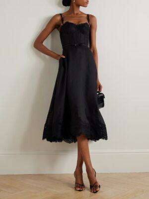 A woman stands against a light-colored wall, wearing a Zimmermann Pleated Tiered Polka-Dot Organza Midi Dress with a fitted bodice and lace trim, black heeled sandals, and holding a small black handbag.