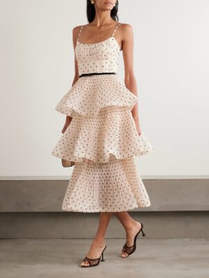 A woman wearing a sleeveless, light pink tiered midi dress inspired by the Zimmermann Ascension Floral Ruffled Cotton Midi Dress, with black polka dots, a thin black belt, and black heeled sandals stands against a plain light-colored wall.
