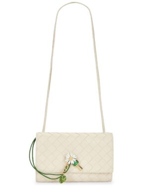 A small cream-colored woven crossbody bag with a long strap, inspired by the Bottega Veneta Embellished Intrecciato-Trimmed Leather Shoulder Bag, featuring a daisy flower and green leaf charm on the front flap.