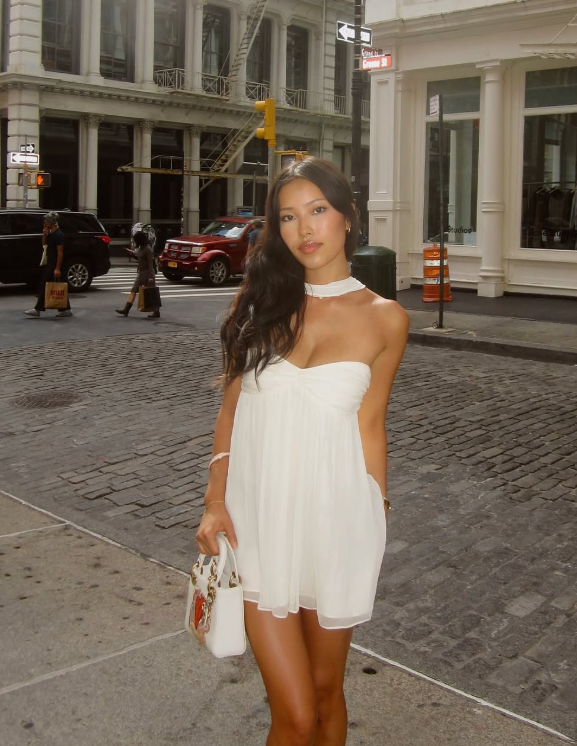 A woman in a strapless white lingerie dress stands on a city sidewalk near a cobblestone street, holding a white handbag and looking at the camera. Buildings, parked cars, and a street intersection are visible in the background.