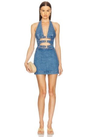 A woman stands facing forward, wearing a sleeveless, halter-neck, blue denim mini dress with cut-out details at the waist and chest, reminiscent of the Cult Gaia Matisse Knit Dress. She holds a beige clutch and wears beige sandals.