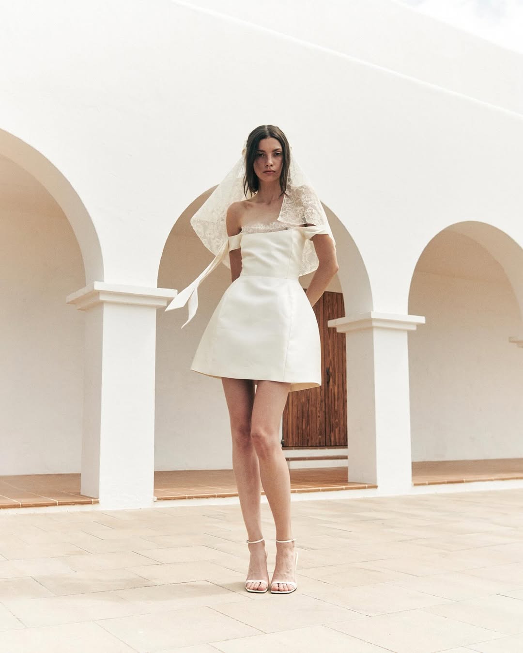 A woman stands outdoors in front of a white building with arches, wearing a short, off-the-shoulder white dress inspired by satin party dresses 2025, featuring decorative lace and a matching headpiece, paired with strappy high-heeled sandals.