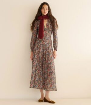 A woman stands against a plain light-colored wall, wearing a long-sleeved, ankle-length Dôen Orphelia Dress with floral print, a red scarf around her neck, and brown flat shoes. She faces forward with a neutral expression.