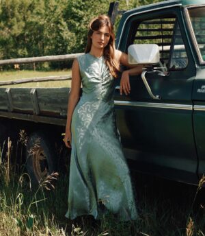 A woman in a light green Dôen Belline Dress stands next to a dark green pickup truck in a grassy outdoor setting, with a wooden fence and trees in the background.