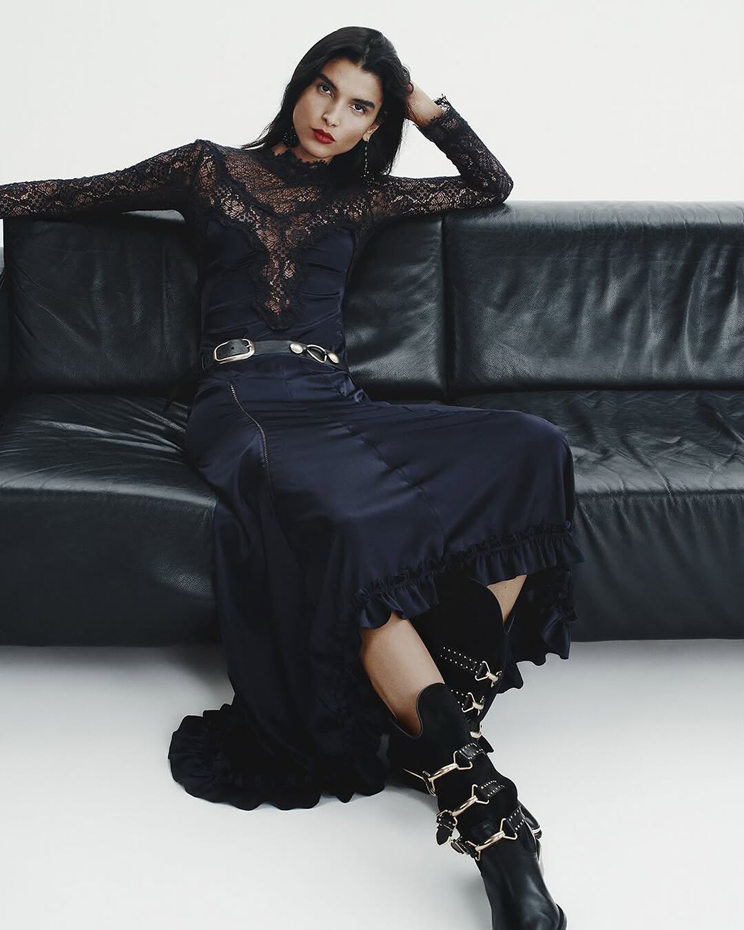 A person with long dark hair sits on a black leather couch, wearing a black lace top, a long skirt with ruffle details inspired by satin party dresses 2025, and black boots adorned with decorative gold chains.