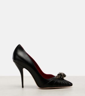 A black high-heeled pump with a pointed toe, featuring pleated detailing and a metallic skull embellishment on the front. Inspired by the Valentino Rockstud 65 Leopard-Print Calf Hair Pumps, it has a red interior lining and a thin stiletto heel.