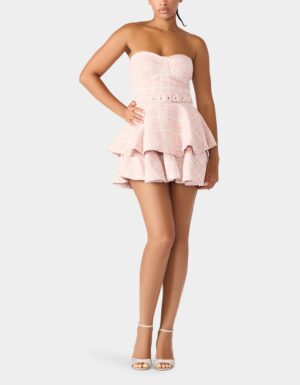 A woman wears a Betsey Johnson Belted Tweed Dress, featuring a strapless, light pink textured mini design with a belted waist and tiered ruffle skirt. She stands against a plain light gray background in white high-heeled sandals.