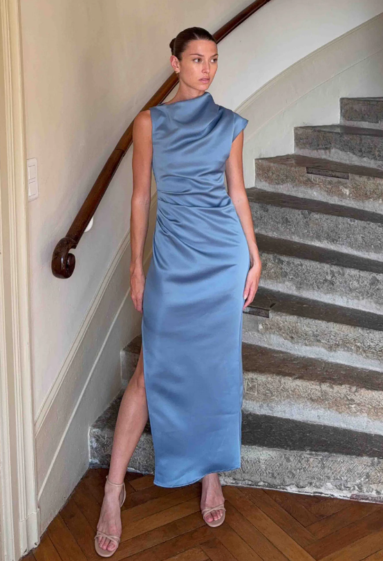 A woman stands on an indoor stone staircase wearing a sleeveless, light blue satin dress—an elegant choice for what to wear to a fall wedding. Her hair is pulled back, complemented by nude open-toe heels and a high neckline with a side slit.