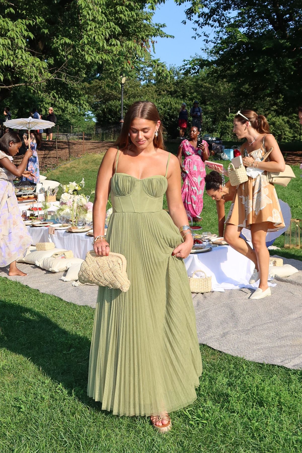 A woman in a light green pleated dress—perfect inspiration for What to Wear to a Fall Wedding—stands on grass at an outdoor picnic event, holding a woven clutch. Others mingle near picnic setups with plates, cushions, and flowers among surrounding trees.