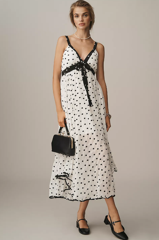 A person stands against a plain background wearing a sleeveless white maxi slip dress styled for Halloween, featuring black polka dots, black trim, and a black bow at the bust. They hold a black handbag and wear black shoes and a pearl necklace.