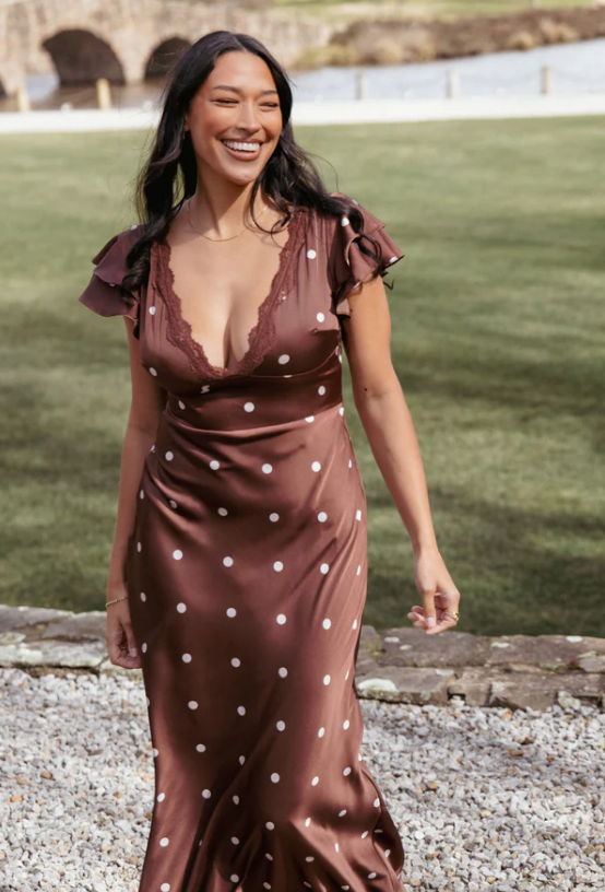 A woman stands outdoors on a stone path, wearing a brown dress with white polka dots and lace trimโan ideal choice for Everyday Dresses for Fall. She has long dark hair, is smiling, and a grassy area is visible in the background.