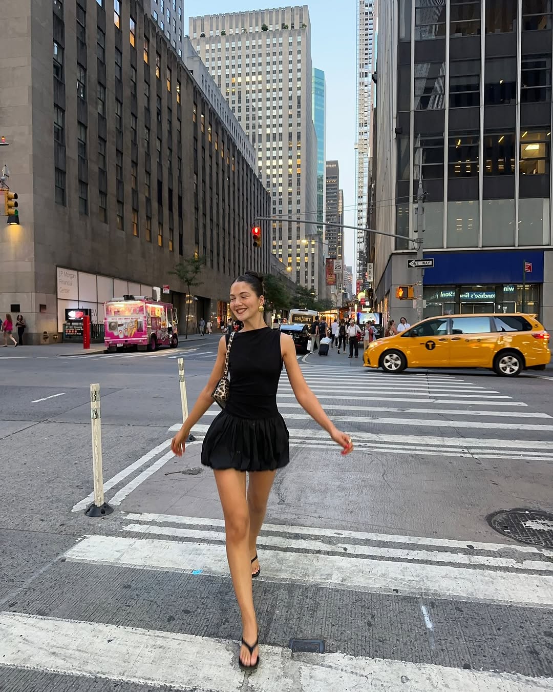 A woman in a minimalist black dress under $150 and sandals walks across a city crosswalk, surrounded by modern silhouettes of tall buildings, a yellow taxi, and bustling people in the background.