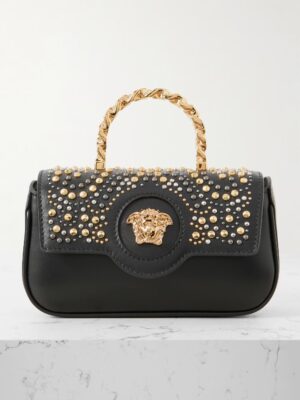A Versace Embellished Leather Tote featuring gold and silver studs, a gold Medusa head emblem at the center, and a twisted gold handle, placed on a white marble surface against a light background.