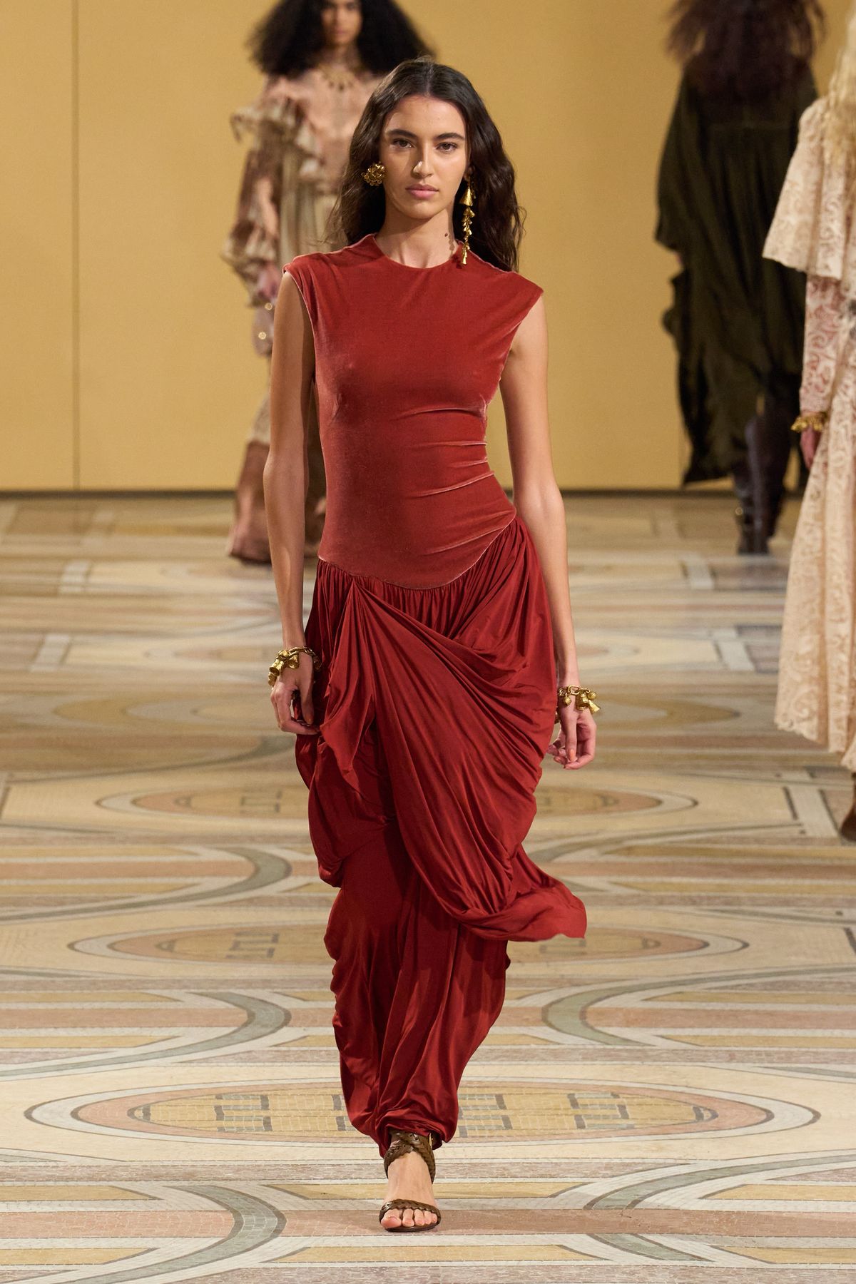 A model walks a runway wearing a sleeveless, rust-colored velvet top paired with a matching draped, gathered skirt and sandals—an inspiring look for What to Wear to a Fall Wedding—set against a beige background with other models in the distance.
