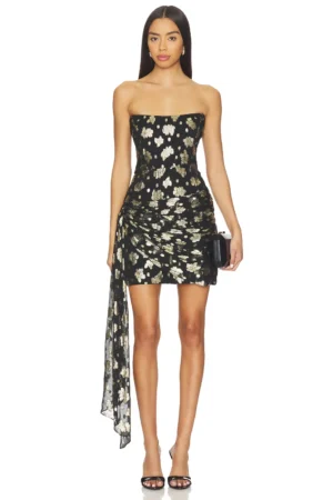 A woman stands facing forward, wearing the Amanda Uprichard Avalee Strapless Skort Romper with a gold and white floral pattern, a side drape detail, black open-toe heels, and holding a small black clutch.