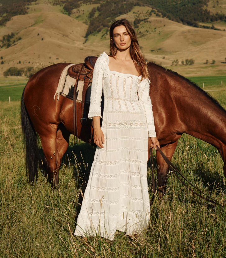 A woman in a flowing boho maxi dress stands in a grassy field, holding the reins of a saddled brown horse. Rolling hills and trees stretch into the distance—an idyllic scene perfect for weekend getaways under the clear sky.