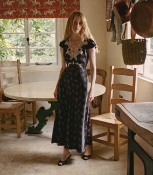 A woman in a long, dark Coraline dress with a white lace collar stands in a rustic kitchen with wooden chairs, a round table, and hanging pots. Sunlight filters through a window with a red horse-patterned valance.