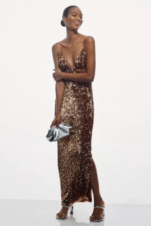 A woman wearing a bronze sequin sleeveless gown with thin straps, reminiscent of the Endless Rose Rosette Knit Midi Dress, stands against a white background holding a silver clutch and wearing silver heeled sandals. She smiles with her arms loosely crossed.