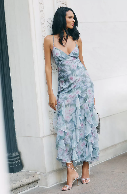 A woman stands against a marble wall wearing a pale blue, floral-patterned dress with thin straps and a ruffled skirt—perfect for layering dresses for autumn. She holds a metallic clutch and wears strappy high-heeled sandals.